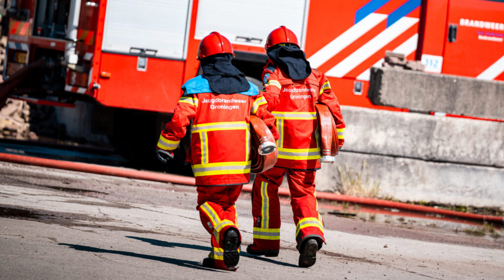 twee jeugdbrandweerlieden die met een brandweerslang richting een brandweerauto lopen.